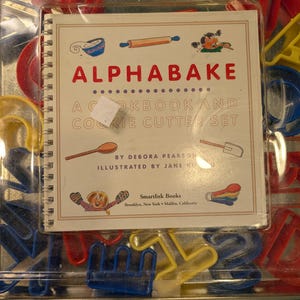 May include: A clear plastic container holding a cookbook and cookie cutter set. The cookbook cover reads "ALPHABAKE COOKBOOK AND COOKIE CUTTER SET" with illustrations. Various colourful cookie cutters in red, yellow, and blue are visible.