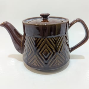 Può includere: Una teiera in ceramica marrone con un motivo geometrico. La teiera ha un coperchio e un manico.