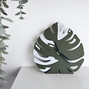Puede incluir: Un reloj decorativo con forma de hoja de monstera. La esfera del reloj es verde oscuro con detalles blancos y manecillas negras. El reloj está sobre una superficie blanca contra una pared blanca. Una planta colgante está en la esquina superior izquierda.