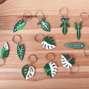 Puede incluir: Un conjunto de 10 llaveros con diferentes diseños de hojas verdes y blancas. Cada llavero tiene un anillo plateado.