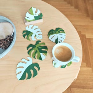Può includere: Set di sei sottobicchieri a forma di foglia di monstera, di colore verde e bianco. I sottobicchieri sono realizzati in un materiale spesso e resistente, e sono perfetti per proteggere il tuo tavolo dal calore e dai graffi.