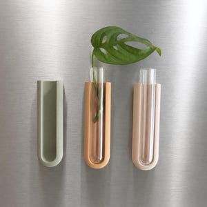 Op de afbeelding: Drie aan de muur gemonteerde vaasjes op een rij. De vazen zijn in groen, bruin en roze tinten. Elke vaas bevat een heldere glazen buis. Eén vaas bevat een groen blad. De vazen zijn op een zilveren oppervlak gemonteerd.