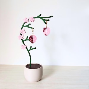 Könnte beinhalten: Ein rosa-grüner, blattartiger Ohrringständer mit rosa Blüten. Der Ständer ist aus Holz und hat einen Topf am Boden.