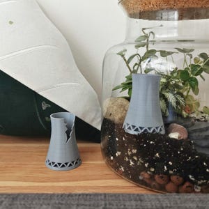 Puede incluir: Dos modelos en miniatura grises de torres de refrigeración, uno dañado, sobre una superficie de madera. Uno está dentro de un terrario de vidrio con plantas y rocas. Los modelos tienen un diseño geométrico con recortes triangulares en la base.