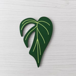 Peut inclure: Découpe en bois en forme de feuille vert foncé avec des veines vert clair. La découpe est une représentation stylisée d'une feuille de plante, avec une finition peinte lisse. La feuille est posée sur un fond en bois blanc.