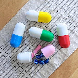 May include: A collection of colorful, capsule-shaped containers. One is open, revealing pink items and blue candies. The capsules are white with ends in yellow, green, red, and blue. The items are arranged on a patterned cloth.