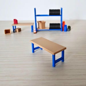 May include: A miniature workshop scene. A tan table with blue legs is in the foreground. Behind it, a blue shelving unit holds black barrels and a cardboard box. Additional small tan tables, boxes, and containers are also present.