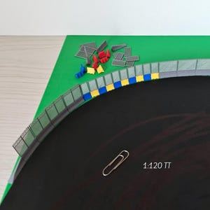 Può includere: Un modello in miniatura di pista da corsa con una superficie in asfalto nero, un campo interno verde e una recinzione grigia. La pista ha segni di cordolo blu e giallo. Piccoli accessori colorati sono sparsi sulla superficie verde. Sono visibili anche una graffetta e il testo "1:120 TT".