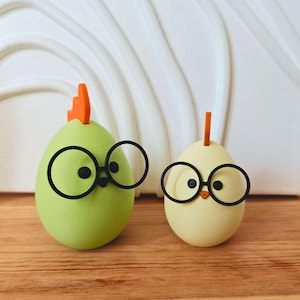 Può includere: Due piccoli pulcini di Pasqua in ceramica con becchi arancioni e occhiali rotondi neri. Il pulcino a sinistra è verde e il pulcino a destra è giallo.