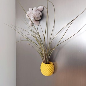 Peut inclure: Un pot de fleur jaune en forme d'ananas avec une surface texturée, contenant une plante verte. Le pot est fixé à une surface métallique. Un petit lapin en peluche est également fixé.