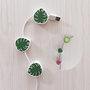 Puede incluir: Organizador de cables blanco con tres clips en forma de hoja con un diseño de hoja verde. El organizador tiene un conector USB en un extremo. Se muestra una versión más pequeña del organizador en un recuadro circular.