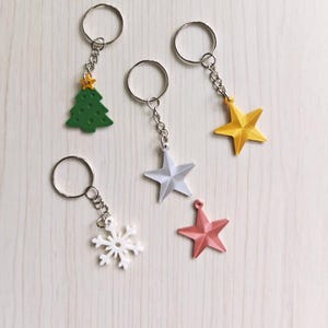 Könnte beinhalten: Eine Sammlung von Schlüsselanhängern zum Thema Weihnachten. Die Schlüsselanhänger zeigen einen grünen Weihnachtsbaum, einen weißen Schneeflocken und drei sternförmige Anhänger in Silber, Gelb und Rosa. Jeder Anhänger ist an einer silbernen Kette und einem Ring befestigt.