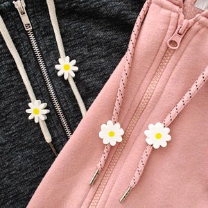 May include: Close-up of two hooded sweatshirts, one charcoal gray and one dusty rose. Both have white drawstrings with daisy-shaped embellishments. The zippers are silver and pink, respectively. The daisy accents have white petals and yellow centers.