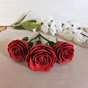 Könnte beinhalten: Drei künstliche rote Rosen mit grünen Stielen und Blättern sind abgebildet. Die Rosen sind der Mittelpunkt, mit weißen Blumen im Hintergrund. Die Rosen haben detaillierte Blütenblätter.
