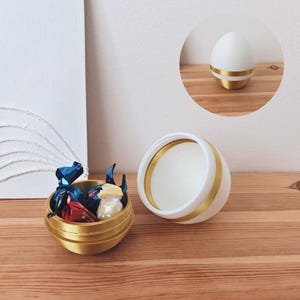 Puede incluir: Un recipiente blanco con forma de huevo con una banda dorada alrededor del medio. El recipiente está abierto y revela un recipiente más pequeño con forma de huevo dorado con una banda dorada alrededor del medio. El recipiente más pequeño está lleno de chocolates envueltos.