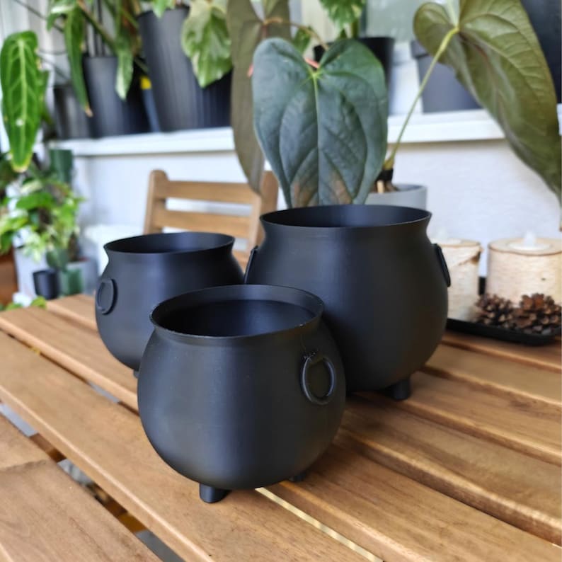 Three matte black 3D-printed cauldron planters of different sizes displayed on a wooden table surrounded by green houseplants