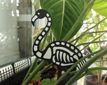 May include: A black and white flamingo skeleton plant stake. The flamingo has a long, curved neck and a detailed skeletal design. It is placed among green plants, adding a unique touch to any indoor or outdoor garden.