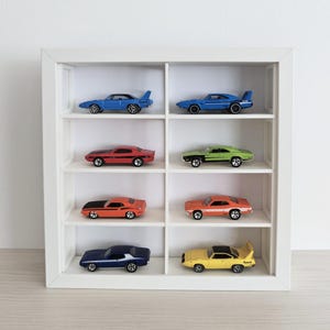 May include: A white square display case with eight individual compartments, each housing a die-cast toy car. The collection includes various models in blue, red, green, orange, and yellow, arranged in two columns across four horizontal shelves.