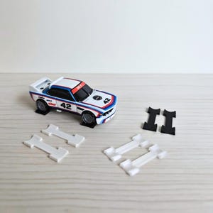 Op de afbeelding: Een wit, blauw en rood racewagenmodel met het nummer 42 op de motorkap en zijkanten. De auto staat op zwarte standaards, met verschillende witte en zwarte plastic onderdelen in de buurt. De auto heeft een racestreepontwerp.