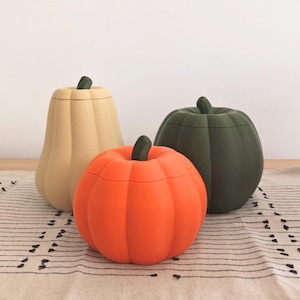 Può includere: Tre contenitori decorativi a forma di zucca con coperchio. Uno è alto e beige, uno è rotondo e arancione brillante, e uno è rotondo e verde bosco scuro. Ogni zucca presenta un piccolo stelo verde come maniglia sopra il coperchio.