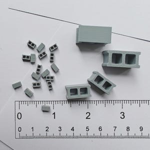 May include: A set of miniature concrete blocks in various sizes, including a stack of three blocks, a single block, and a group of smaller blocks. The blocks are a light gray color and are arranged on a white surface with a ruler for scale.