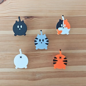 Puede incluir: Cinco adornos con forma de gato, cada uno con un color y un patrón diferentes. Los adornos están hechos de un material suave y flexible y tienen una parte posterior plana. Los gatos están todos mirando hacia adelante con sus colas enroscadas detrás de ellos. Los colores de los gatos son negro, blanco, gris, naranja y calico.