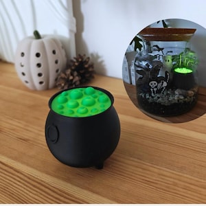 May include: A black cauldron with green, bubbling liquid, a Halloween decoration. The cauldron has a round shape with three small feet. A white pumpkin and a glass jar with a Halloween scene are in the background.