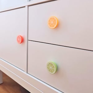 May include: A white cabinet featuring three circular drawer knobs shaped like citrus fruit slices. One knob is orange, one is a lighter orange, and one is a light green, each displaying a detailed segment pattern resembling a sliced fruit.