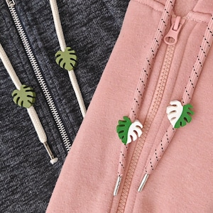 May include: Close-up of two hoodies, one gray and one pink, with decorative leaf-shaped cord ends. The gray hoodie has green leaf accents, while the pink one has green and white leaf accents. The hoodies have white and patterned drawstrings.