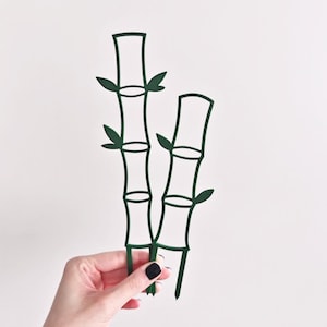 May include: Dark green bamboo plant stakes with leaf accents. The stakes are made of a thin, cut-out material and are designed to support plants. The stakes are held by a person with a white background.