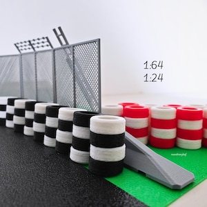 Op de afbeelding: Miniatuurmodel van een racebaan met zwart-wit gestreepte barrières, rood-witte barrières, een grijs hek en een grijze helling. De scène staat op een groen en zwart oppervlak. De tekst "1:64" en "1:24" is zichtbaar.