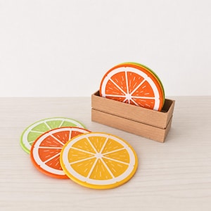 Op de afbeelding: Een set ronde onderzetters met levendige citrusvrucht-ontwerpen in oranje, geel en limoengroen. De onderzetters zijn gemaakt van een flexibel materiaal en worden bewaard in een rechthoekige, lichtgekleurde houten houder met een open bovenkant.