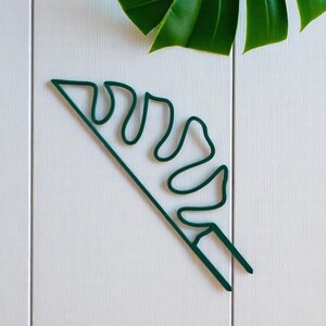Könnte beinhalten: Eine dunkelgrüne Pflanzenstütze aus Metall in Form eines Monstera-Blattes. Die Stütze befindet sich auf einer weißen Holzoberfläche, mit einem echten Monstera-Blatt im Hintergrund. Sie dient dazu, Pflanzen beim Wachsen zu unterstützen.