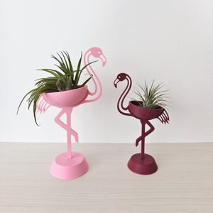 Può includere: Due fioriere a forma di fenicottero, una rosa chiaro e una bordeaux, ciascuna con una piccola pianta. Le fioriere hanno un contenitore a forma di ciotola tenuto dal collo e dal becco del fenicottero, con le zampe del fenicottero che formano un supporto.