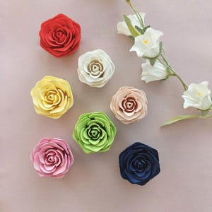 May include: A collection of seven individual rose-shaped decorative items in red, white, yellow, light pink, lime green, beige, and dark blue, accompanied by a single stem of white bell-shaped flowers with green leaves and yellow centers.