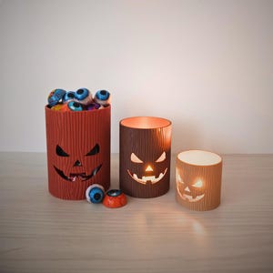 May include: Three cylindrical containers with a wood grain texture and carved jack-o'-lantern faces. The largest container is orange and filled with round, eye-themed candies. The two smaller containers are brown and tan, illuminated from within.