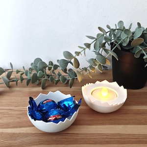 Puede incluir: Dos portavelas de cerámica blanca en forma de huevo. Un portavelas tiene una vela de té encendida en su interior. El otro portavelas está lleno de chocolates envueltos en azul y plata.
