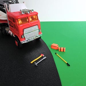 Puede incluir: Un camión de bomberos de juguete rojo con rejilla plateada, ventanas naranjas y tres bocinas plateadas en el techo. Junto al camión hay dos pequeñas cajas de almacenamiento naranjas, un hacha con mango amarillo, una palanca gris y un poste.