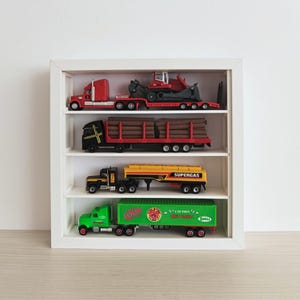 Könnte beinhalten: Eine weiße Vitrine mit vier Regalen, die jeweils einen Spielzeug-LKW enthalten. Die LKWs sind rot, schwarz, gelb und grün. Der grüne LKW trägt das Wort "SOUR". Die Vitrine steht auf einer weißen Oberfläche.
