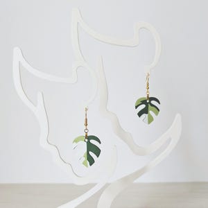 Peut inclure: Une paire de boucles d'oreilles en or avec des breloques en forme de feuilles de monstera vertes et blanches.