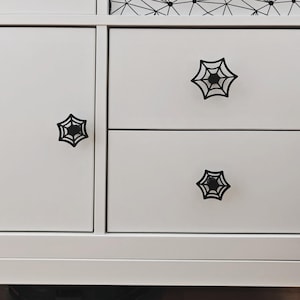 May include: White cabinet with black spiderweb-shaped knobs. The knobs are centered on the cabinet doors and drawers. The cabinet has a clean, modern design, suitable for a Halloween or gothic-themed room.