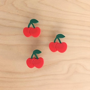 Könnte beinhalten: Drei rote, kirschförmige Dekorationen mit grünen Blättern sind auf einer hellen Holzoberfläche angeordnet. Jede Kirsche hat zwei rote Kugeln und einen grünen Stiel mit einem Blatt.