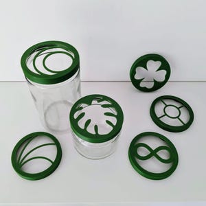 Peut inclure: Des bocaux en verre transparent avec des couvercles décoratifs verts aux motifs variés, dont un trèfle à quatre feuilles et un symbole infini. Les bocaux sont disposés sur une surface blanche, mettant en valeur les motifs complexes des couvercles.