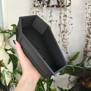 Puede incluir: Una maceta negra mate en forma de ataúd con tres orificios de drenaje. La maceta tiene forma rectangular con lados angulados y adornos decorativos. La maceta se sostiene frente a un fondo de plantas verdes y enredaderas colgantes.