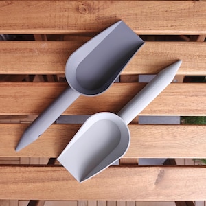 Two grey plastic scoops with long handles are arranged on a wooden surface. One scoop is a darker shade of grey than the other. The scoops have a rounded shape and are designed for scooping materials.
