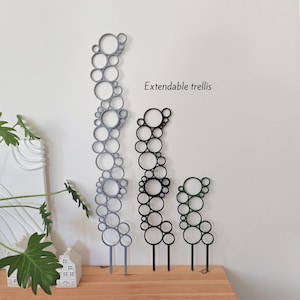 Puede incluir: Tres soportes extensibles para plantas en gris, negro y verde, cada uno con una serie de círculos conectados. Los soportes se muestran sobre una superficie de madera contra una pared blanca. El texto en la pared dice "Extendable trellis."