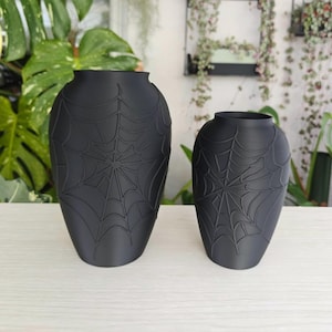 May include: Two matte black vases of different sizes, each featuring a raised, embossed spider web pattern covering the exterior surface. The vases have a rounded, bulbous shape with a narrow opening at the top and a smooth, uniform finish.
