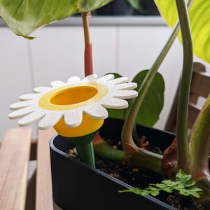 Puede incluir: Un dispositivo de riego en forma de margarita blanca con un centro amarillo y un tallo verde. El dispositivo se inserta en una maceta negra con tierra y una planta verde.