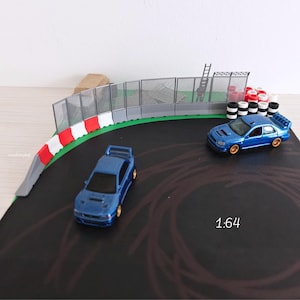 Könnte beinhalten: Zwei blaue Spielzeugautos auf einer Miniatur-Rennstrecke. Die Strecke verfügt über eine grüne und graue Barriere, einen Maschendrahtzaun und Reifenbarrieren. Der Maßstab ist 1:64.