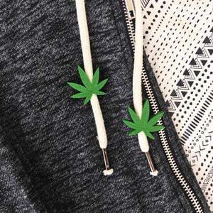 May include: Close-up of a dark gray hoodie with white drawstrings and two green cannabis leaf charms. The zipper is silver, and a black and white patterned fabric is visible in the background.